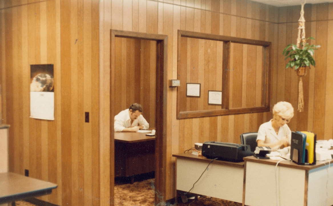 1980 photo of a wood paneled lobby with a woman sitting at the front desk and a man sitting at his office desk