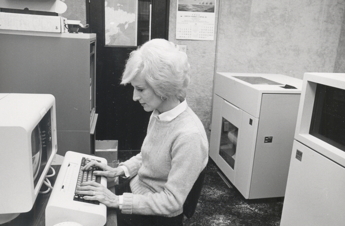 Black and white photo of a blonde woman typing on a large desktop computer