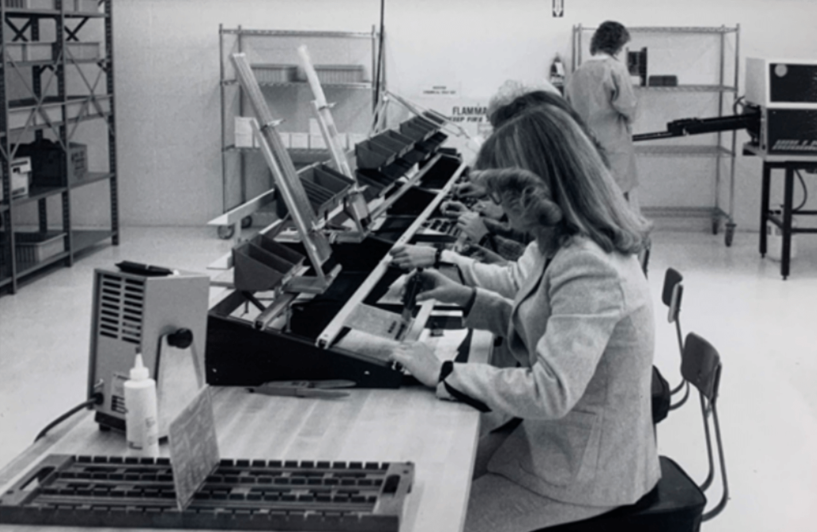 Black and white photo of women sitting on a production line placing components on a printed circuit board