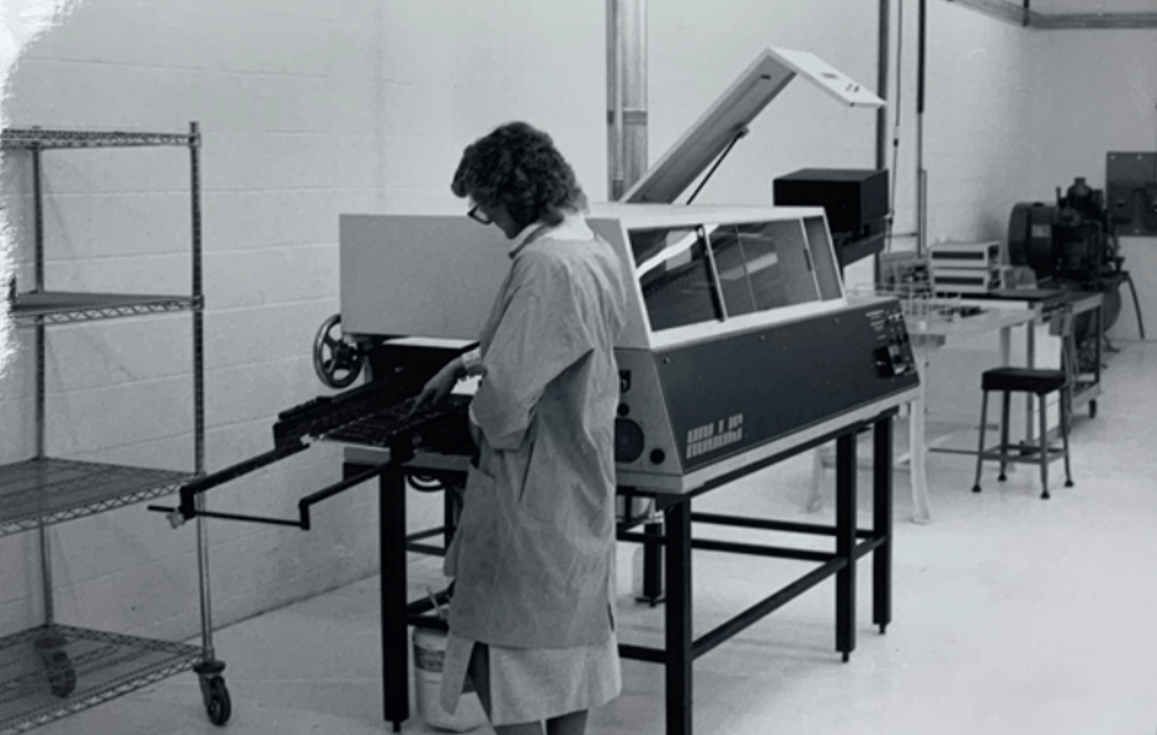 Black and white photo of a woman standing in front of a wave solder machine